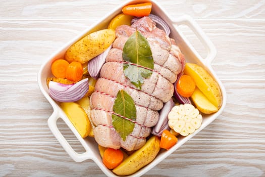 Raw wrapped rolled sliced pork in white casserole dish with potatoes, vegetables and herbs on rustic white wooden background top view. Pork roll with vegetables ready to be prepared.