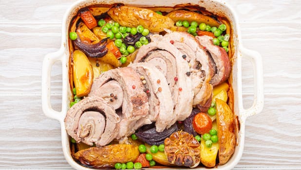 Rolled sliced pork roasted in white casserole dish with potatoes, vegetables and herbs on rustic white wooden background top view. Baked pork roll with vegetables for dinner.