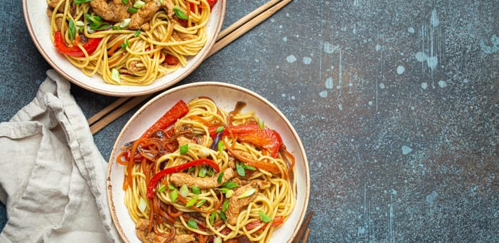 Two bowls with Chow Mein or Lo Mein, traditional Chinese stir fry noodles with meat and vegetables, served with chopsticks top view on rustic blue concrete background, space for text.