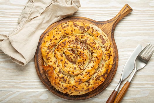 Burek made of filo dough with filling on cutting board, white wooden rustic background top view. Traditional savoury spiral pie of Balkans, Middle East and Central Asia.