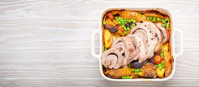 Rolled sliced pork roasted in white casserole dish with potatoes, vegetables and herbs on rustic white wooden background top view. Baked pork roll with vegetables for dinner.