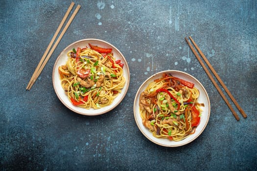 Two bowls with Chow Mein or Lo Mein, traditional Chinese stir fry noodles with meat and vegetables, served with chopsticks top view on rustic blue concrete background.