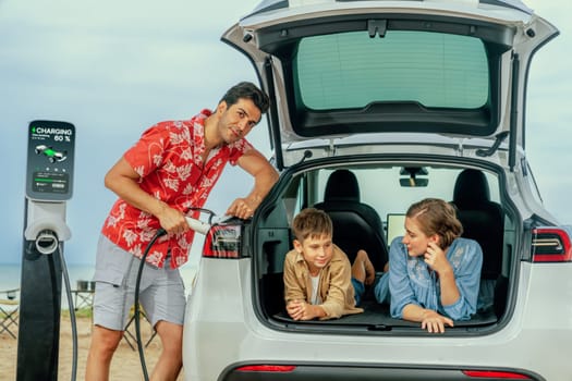 Family vacation trip traveling by the beach with electric car, lovely family sit on the trunk, charging EV car battery with green and sustainable energy. Family travel and eco-friendly car. Perpetual