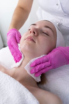Beauty Specialist Cleans Woman's Face, Neck Skin Using Cotton Pads Before Beauty Procedure in Spa Salon. Cleaning Skin With Micellar Water. Young Female Enjoying Facial Skincare. Vertical.