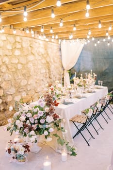 Bouquets of flowers and lit candles stand on a festive table on the terrace in the light of garlands. High quality photo