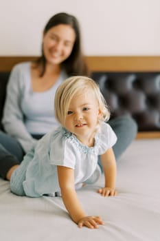Little girl crawls on the bed against the background of a smiling mother. High quality photo