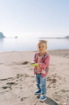 Little girl stands on the beach with a toy shovel full of sand. High quality photo