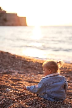 Little girl sits on the seashore and sorts through the pebbles with her hand. Back view. High quality photo