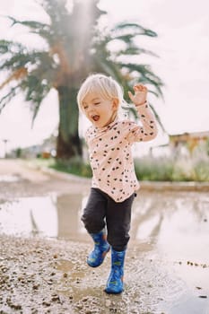 Smiling little girl in rubber boots jumping on a puddle. High quality photo