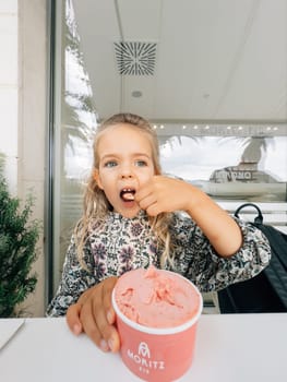 Little girl eats pink ice cream from a cup with a spatula at the table. Caption: Moritz Eis. High quality photo