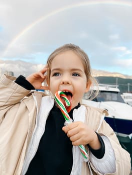 Little girl licks a lollipop against the backdrop of a rainbow over the mountains. High quality photo