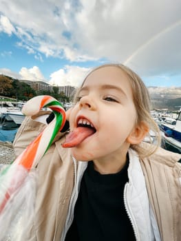 Little girl with her tongue hanging out reaches for a lollipop against the backdrop of a rainbow. High quality photo