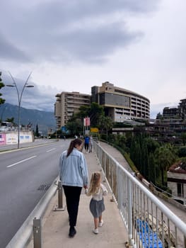 Mom and little girl walk along the sidewalk along the highway to high-rise buildings. Back view. High quality photo