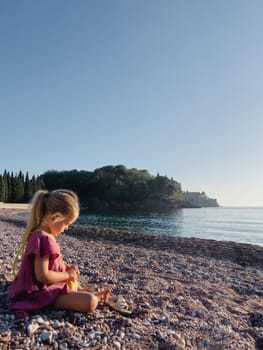 Little girl is sitting on a pebble beach by the sea and opening a small bag. High quality photo