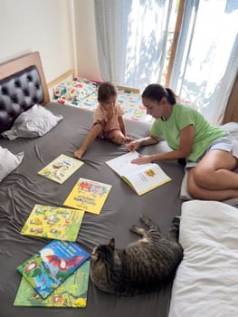 Mom reads a little girl bright books lying on the bed next to a tabby cat. High quality photo