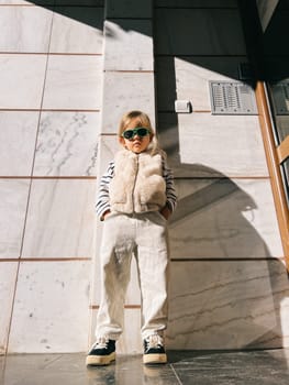 Little girl in sunglasses with her hands in her pants pockets stands near the wall. High quality photo