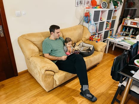 Dad reads a book to a little girl sitting on the couch next to a lying cat. High quality photo