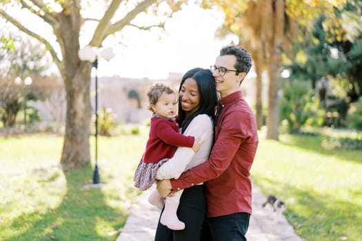 Smiling dad hugging mom from behind with a little girl in her arms in the sunny park. High quality photo