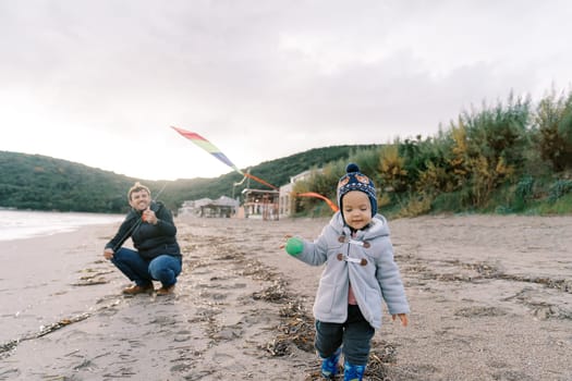 Little girl walks along the beach with her dad squatting with a kite in the background. High quality photo