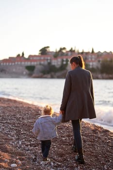 Mom and a little girl walk holding hands along the seashore to the island of Sveti Stefan. Back view. High quality photo