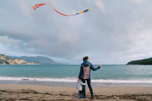 Small child hugs his mother leg standing by the sea with a kite. Back view. High quality photo