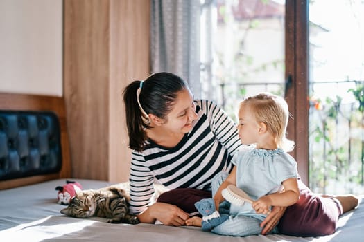 Smiling mother sitting on bed near little girl holding hair brush. High quality photo