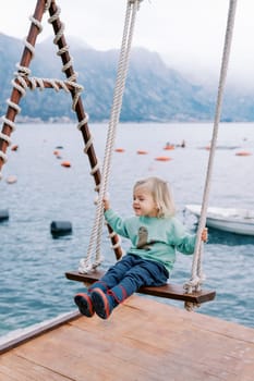 Little smiling girl swings on a swing on a pier by the sea. High quality photo