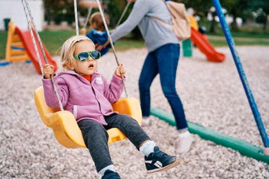 Little girl in sunglasses swings on a swing in the playground. High quality photo