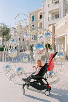 Little girl in a stroller sits near the New Year installation in the form of a Christmas ball. High quality photo