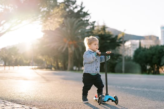 Little girl stands holding the steering wheel of a scooter on the road and looks ahead. High quality photo