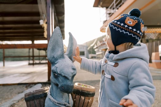 Little girl stands near a sculpture of a donkey and strokes his ears. High quality photo