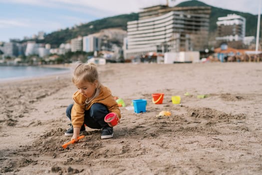 Little girl on the beach pours sand with a toy shovel into a plastic bucket while squatting. High quality photo