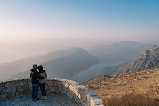 Couple of travelers in jackets hug on an observation deck over the Bay of Kotor in the fog. Montenegro. Back view. High quality photo