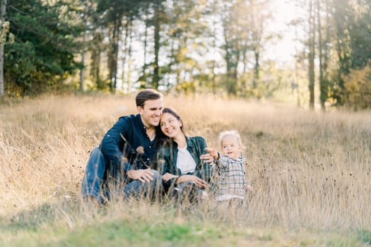 Little girl sits on the lawn next to hugging mom and dad. High quality photo