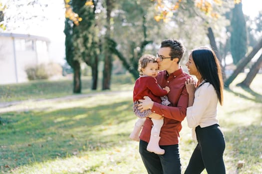 Smiling mom hugging shoulders of dad kissing little girl in his arms. High quality photo