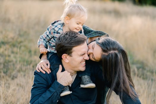 Mom hugs dad with a little girl on his shoulders from behind and leaning over kisses his cheek. High quality photo