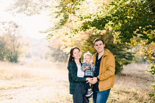 Smiling mom and dad with a little girl in their arms stand on the sunny edge of the forest. High quality photo