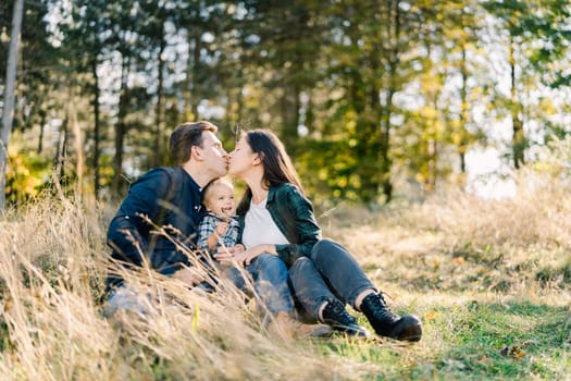 Dad and mom kiss while sitting with a little girl in a clearing near the forest. High quality photo