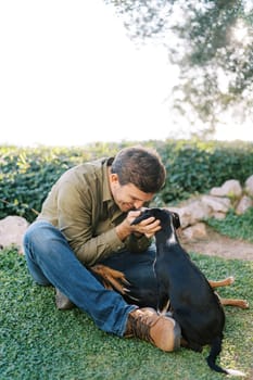 Smiling young man sits on the lawn and touches with his nose the nose of a big puppy. High quality photo