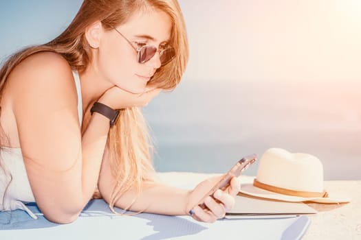 Successful business woman in yellow hat working on laptop by the sea. Pretty lady typing on computer at summer day outdoors. Freelance, travel and holidays concept.