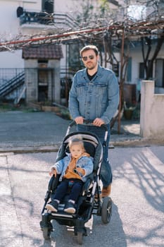 Dad with a little girl in a stroller walks along the road near the house. High quality photo