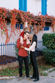 Mom hugs a smiling dad with a little girl on his shoulders while standing in the courtyard of the house. High quality photo