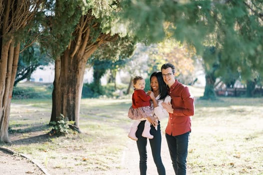 Smiling dad hugging mom from behind with a little girl in her arms near a tree. High quality photo