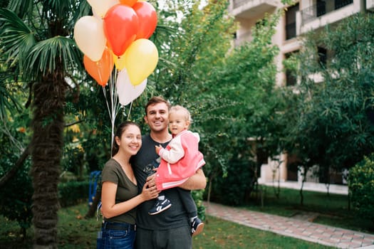 Mom hugs dad with a little girl in his arms in the garden with colorful balloons. High quality photo