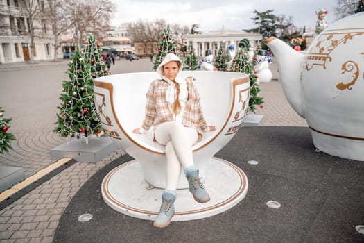 Woman Christmas Square. She sits in a large white cup, dressed in a light suit. With trees decorated with Christmas tinsel in the background.