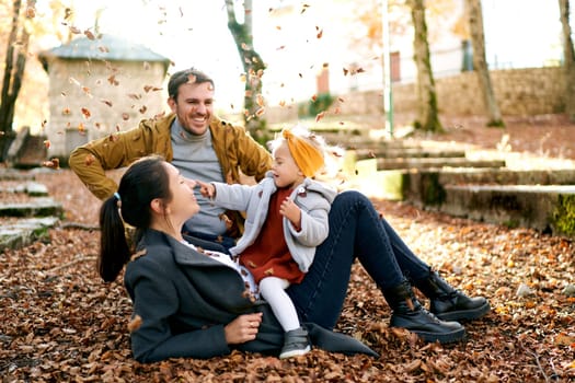 Laughing dad sits next to mom lying on the ground with a little girl on her stomach under falling leaves. High quality photo