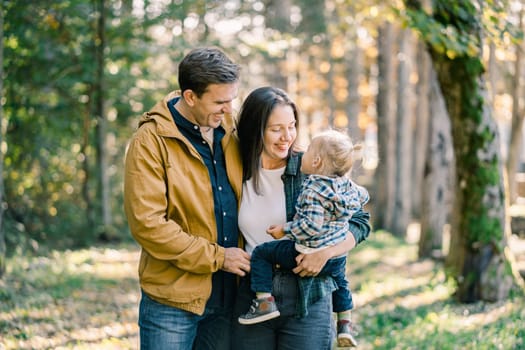 Smiling dad hugging mom with little daughter in her arms in park. High quality photo