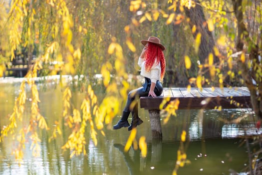 Autumn lake woman. In autumn, she sits by the pond on a wooden pier and admires nature with red hair and a hat. Tourism concept, weekend outside the city