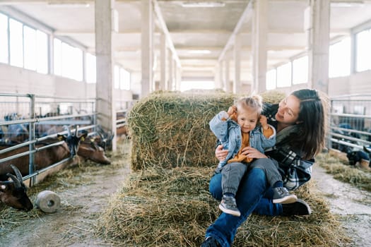 Smiling mom with little girl on her lap sitting on haystack near goat pens at farm. High quality photo