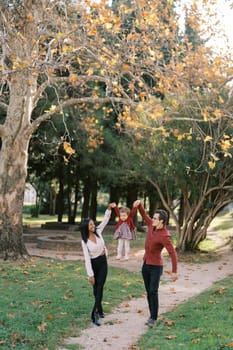 Mom and dad are standing on the path in the park, holding their little daughter in their arms raised. High quality photo